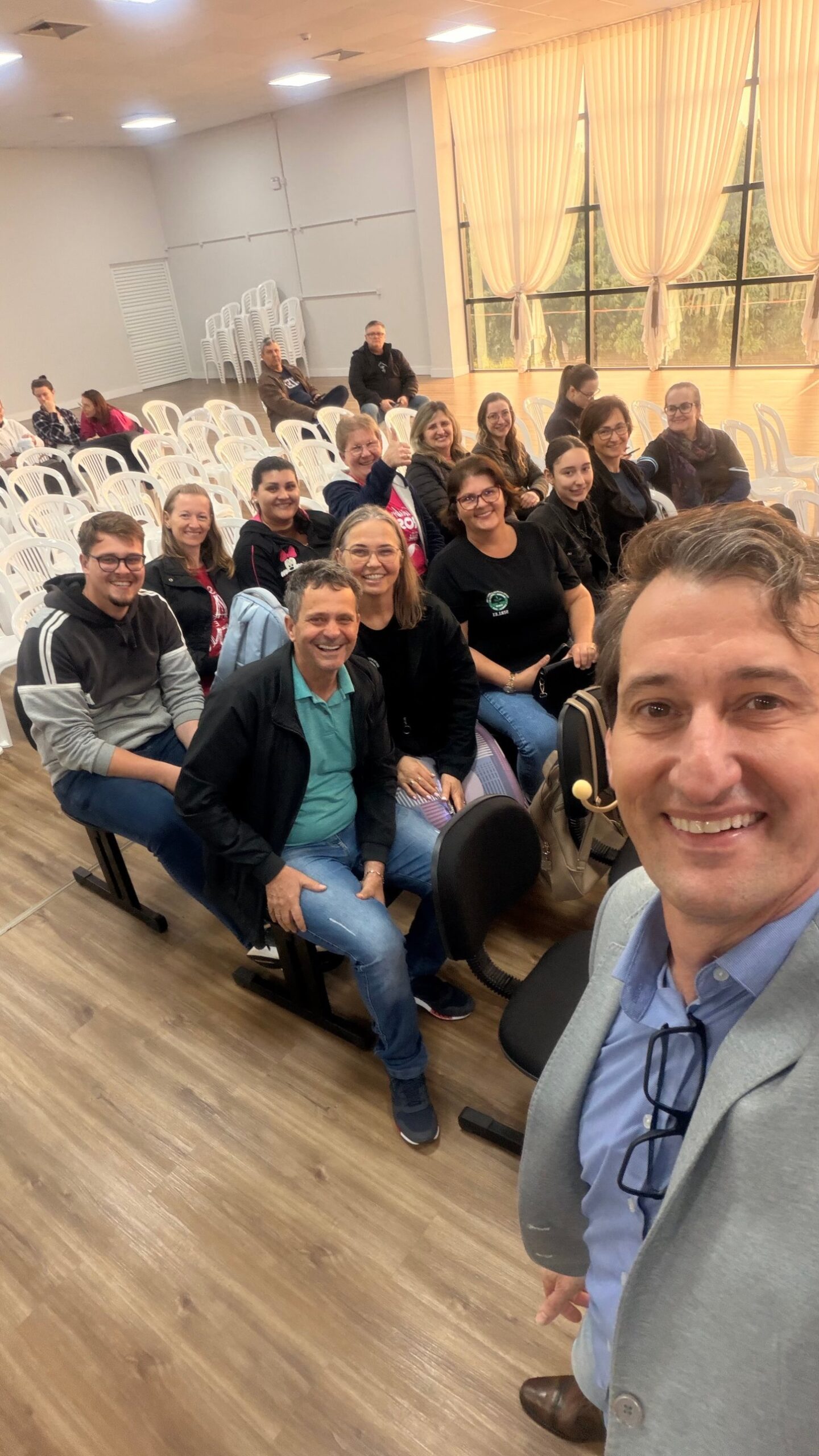 Edney Prigol com um grupo de servidores da educação de Iporã do Oeste sorrindo nas primeiras fileiras, com cortinas e janelas ao fundo